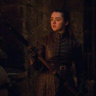 an image of Arya Stark from HBO's game of thrones series, season eight. Here, she is holding a weapon as light from a fire in the distance brings light to her face.